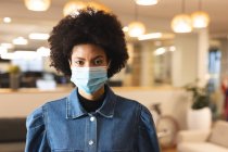 Porträt einer kreativen Frau gemischter Rasse mit Gesichtsmaske bei der Arbeit, die in die Kamera blickt. Arbeit im kreativen Geschäft in einem modernen Büro während der Coronavirus-Pandemie. — Stockfoto
