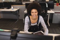 Sonriente mezcla de raza femenina creativa en el trabajo, sentado en el escritorio, utilizando la computadora. trabajar en un negocio creativo en una oficina moderna. - foto de stock
