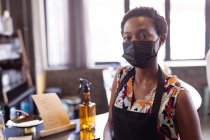 Portrait de femme afro-américaine portant tablier et masque debout à la distillerie de gin. distillerie de gin artisanale indépendante pendant le concept pandémique covid-19 — Photo de stock