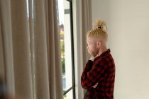 Réfléchi albinos homme afro-américain avec dreadlocks regardant par la fenêtre. télétravail utilisant la technologie à la maison. — Photo de stock
