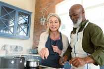 Felice anziano coppia diversificata in cucina indossando grembiuli, cucinare insieme. stile di vita sano e attivo pensionamento a casa. — Foto stock