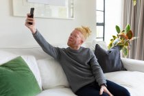 Homme afro-américain albinos dans le salon prenant selfie. loisirs utilisant la technologie, détente à la maison. — Photo de stock