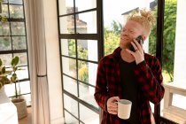 Sorrindo albino homem americano africano com dreadlocks usando smartphone e beber café. trabalho remoto usando a tecnologia em casa. — Fotografia de Stock