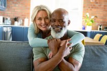 Portrait of happy senior diverse couple in living room sitting on sofa, embracing and smiling. retirement lifestyle, spending time at home. — Stock Photo