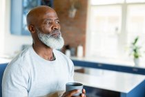 Nachdenklicher älterer afrikanisch-amerikanischer Mann in der Küche, Tasse haltend, wegschauend. Lebensstil im Ruhestand, Zeit zu Hause verbringen. — Stockfoto