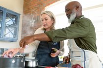 Heureux couple diversifié senior dans la cuisine portant des tabliers, cuisiner ensemble. mode de vie sain et actif à la retraite à la maison. — Photo de stock
