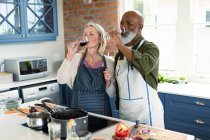 Happy senior diverse couple in kitchen wearing aprons, cooking together, drinking wine. healthy, active retirement lifestyle at home. — Stock Photo