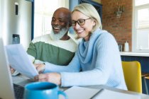 Happy senior diverse couple in kitchen sitting at table, using laptop. retirement lifestyle, at home with technology. — Stock Photo