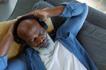 Rilassato uomo anziano afroamericano in soggiorno sdraiato sul divano, con le cuffie. stile di vita di pensione, a casa con la tecnologia. — Foto stock