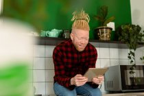 Feliz albino hombre afroamericano con rastas trabajando desde casa y usando tableta. trabajo remoto utilizando tecnología en el hogar. - foto de stock