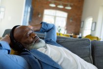 Entspannter älterer afrikanisch-amerikanischer Mann im Wohnzimmer, auf dem Sofa liegend, Kopfhörer tragend. Lebensstil im Ruhestand, zu Hause mit Technologie. — Stockfoto