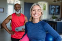 Ritratto di felice coppia anziana diversificata in abiti da ginnastica praticare yoga, guardando la fotocamera. stile di vita sano e attivo pensionamento a casa. — Foto stock