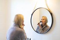 Glückliche ältere kaukasische Frau im Badezimmer, den Spiegel anschauend, sich schminkend. Lebensstil im Ruhestand, Zeit zu Hause verbringen. — Stockfoto