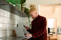 Albino-amerikanischer Mann mit Dreadlocks, der von zu Hause aus arbeitet und Laptop und Smartphone benutzt. Fernbedienung mit Technologie zu Hause. — Stockfoto