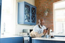 Glücklicher älterer afrikanischer amerikanischer Mann in der Küche mit Smartphone. Lebensstil im Ruhestand, zu Hause mit Technologie. — Stockfoto
