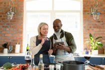 Щаслива старша різноманітна пара на кухні носить фартухи, готуючи разом, використовуючи планшет. здоровий, активний спосіб життя на пенсії вдома . — стокове фото