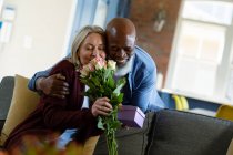 Felice anziano coppia diversificata in soggiorno seduto sul divano, dando fiori e regali. stile di vita di pensione, trascorrere del tempo a casa. — Foto stock