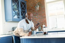 Щасливий старший афроамериканський чоловік на кухні за допомогою смартфона. пенсійний спосіб життя, вдома з технологіями . — стокове фото