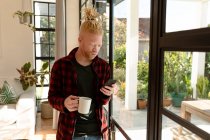 Nachdenklicher Albino-Afrikaner mit Dreadlocks, der Smartphone benutzt und Kaffee trinkt. Fernbedienung mit Technologie zu Hause. — Stockfoto