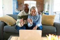 Счастливая пожилая пара в гостиной сидит на диване, пользуется ноутбуком, делает видеозвонок. уход на пенсию образ жизни, дома с технологиями. — стоковое фото