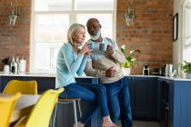 Felice anziano coppia diversificata in cucina seduta al piano di lavoro, bere caffè. stile di vita di pensione, trascorrere del tempo a casa. — Foto stock