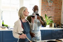 Счастливая пожилая пара на кухне в фартуках, готовящая вместе, пьющая вино. здоровый, активный образ жизни на дому. — стоковое фото