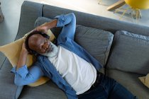 Расслабленный старший африканский американец в гостиной, лежащий на диване, в наушниках. уход на пенсию образ жизни, дома с технологиями. — стоковое фото