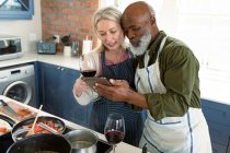 Glückliches älteres Ehepaar in der Küche, Schürzen tragend, gemeinsam kochend, mit Tablet. gesunder, aktiver Lebensstil im Ruhestand zu Hause. — Stockfoto