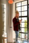 Pensativo albino hombre afroamericano con rastas usando teléfono inteligente y beber café. trabajo remoto utilizando tecnología en el hogar. - foto de stock