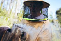 Кавказький старший чоловік у формі бджоляра тримає інструмент з димом, щоб заспокоїти бджіл. Виробництво бджолиного, апіарного та медового виробництва.. — стокове фото