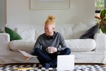 Heureux homme afro-américain albinos assis sur le sol et travaillant de la maison à l'aide d'un ordinateur portable. télétravail utilisant la technologie à la maison. — Photo de stock