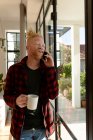 Sorridente uomo afroamericano albino con dreadlocks utilizzando smartphone e bere caffè. lavoro a distanza utilizzando la tecnologia a casa. — Foto stock