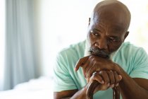 Pensativo hombre afroamericano mayor en el dormitorio sosteniendo bastón. estilo de vida de jubilación, pasar tiempo en casa. - foto de stock