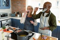 Happy senior diverse couple in kitchen wearing aprons, cooking together, drinking wine. healthy, active retirement lifestyle at home. — Stock Photo