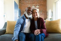 Retrato de feliz casal diverso sênior na sala de estar sentado no sofá, abraçando e sorrindo. estilo de vida da aposentadoria, passar tempo em casa. — Fotografia de Stock