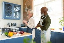 Felice anziano coppia diversificata in cucina cucinare insieme, indossando grembiule. stile di vita sano e attivo pensionamento a casa. — Foto stock
