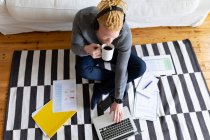 Albino hombre afroamericano sentado en el suelo y trabajando desde casa usando el ordenador portátil. trabajo remoto utilizando tecnología en el hogar. - foto de stock