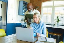 Stressed senior diverse couple in kitchen sitting at table, using laptop. retirement lifestyle, at home with technology. — Stock Photo