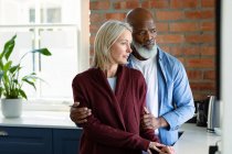 Worried senior diverse couple in kitchen leaning on countertop, embracing and looking away. retirement lifestyle, spending time at home. — Stock Photo