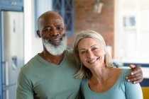 Ritratto di felice coppia anziana diversificata in cucina abbracciando e sorridendo. stile di vita di pensione, trascorrere del tempo a casa. — Foto stock