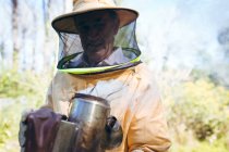 Старший белый мужчина в пчеловодческой форме держит инструмент с дымом, чтобы успокоить пчёл. пчеловодство, пасека и мёд. — стоковое фото