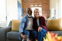 Ritratto di felice coppia anziana diversificata in soggiorno seduta sul divano, abbracciata e sorridente. stile di vita di pensione, trascorrere del tempo a casa. — Foto stock