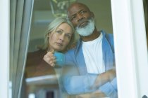 Preoccupato senior coppia diversificata guardando attraverso la finestra e abbracciando. stile di vita di pensione, trascorrere del tempo a casa. — Foto stock