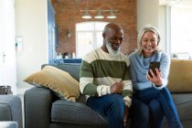 Щаслива старша різноманітна пара у вітальні сидить на дивані, використовуючи смартфон. пенсійний спосіб життя, вдома з технологіями . — стокове фото