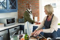 Feliz casal diversificado sênior na cozinha vestindo aventais, cozinhar juntos. estilo de vida saudável e ativo em casa. — Fotografia de Stock