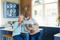 Felice anziano coppia diversificata in cucina seduta al piano di lavoro, bere caffè. stile di vita di pensione, trascorrere del tempo a casa. — Foto stock