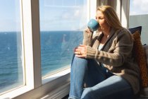 Entspannte kaukasische reife Frau, die Kaffee trinkt und durch das Fenster schaut. Freizeit zu Hause genießen. — Stockfoto