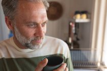 Homme mûr caucasien relaxant buvant du café dans la cuisine et regardant par la fenêtre. profiter du temps libre à la maison. — Photo de stock