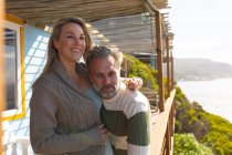 Feliz pareja caucásica de pie en una terraza junto al mar. disfrutar del tiempo libre en la casa frente a la playa. - foto de stock