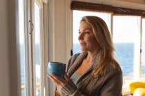 Relaxing caucasian mature woman drinking coffee in the kitchen and looking trough the window. enjoying leisure time at home. — Stock Photo